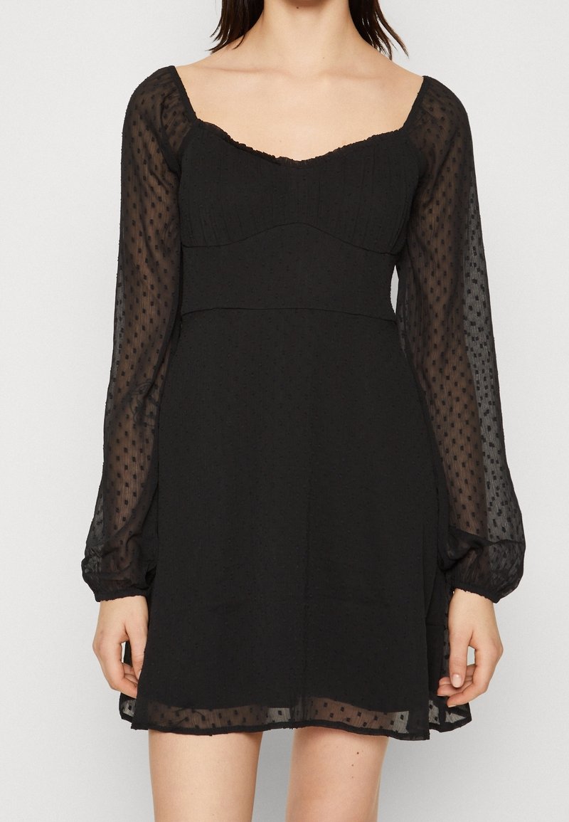 Svart klänning med genomskinliga, puffiga långa ärmar, textilerad tyg och en figurnära liv. Har en fyrkantig halsringning och en lagerpå-lager kjol.