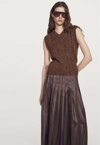 Brun, ärmlös stickad topp med V-ringning; matchad med en lång, plisserad, glänsande, chokladbrun kjol. Modell som bär mörka solglasögon.