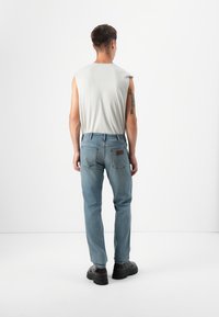 Een persoon staat met de rug naar de kijker, gekleed in een lichtgrijs mouwloos shirt en blauwe jeans, tegen een minimalistische witte achtergrond.
