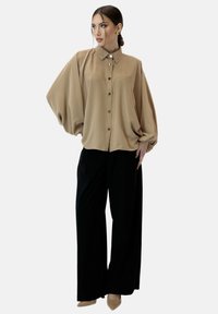 Beige zijden blouse met een knoopsluiting aan de voorkant en luchtige mouwen, gecombineerd met ruime zwarte broeken en beige hakken. Soepele textuur, minimalistisch ontwerp.