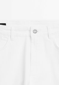 Jean blanc avec fermeture à boutons sur le devant, passants pour ceinture et deux poches avant. Le tissu a une texture lisse.