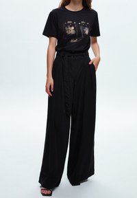 Czarny t-shirt z grafiką i złotym nadrukiem przedstawiającym abstrakcyjne wzory; połączony z wysokim stanem i szerokimi nogawkami czarnych spodni. Całość dopełniają płaskie sandały.