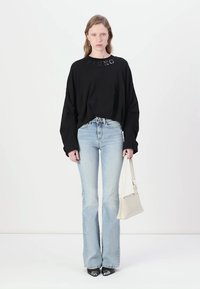 Camicia nera oversize a maniche lunghe con logo "PINKO", jeans a campana blu chiaro e décolleté neri con punta a pigra, mentre tiene una piccola borsa bianca.