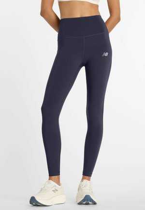 Frau trägt marineblaue Leggings mit hoher Taille, einem kleinen New Balance-Logo am linken Oberschenkel und weißen Sneakern vor weißem Hintergrund.