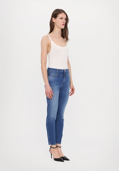 Smale jeans i medium blå denim, som har en klassisk fem-lommers design, lett falming, og ankellang kant, kombinert med en hvit tanktop og svarte hæler.