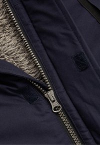 Gros plan sur la fermeture éclair d'une veste marine, partiellement ouverte pour montrer la doublure en polaire grise et les patches Velcro cousus sur les couches de tissu.