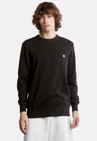Maglione nero a maglia con colletto girocollo, polsini e orlo a coste, con un piccolo logo bianco sul petto, indossato sopra pantaloni bianchi.