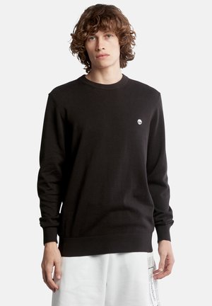 Maglione nero a maglia con colletto girocollo, polsini e orlo a coste, con un piccolo logo bianco sul petto, indossato sopra pantaloni bianchi.