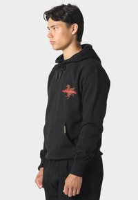 Sudadera negra hecha de una mezcla de algodón, que cuenta con un bolsillo frontal, capucha con cordón y un diseño gráfico rojo en el lado izquierdo del pecho.