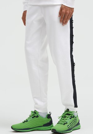 Person trägt weiße Jogginghosen mit schwarzen Seitenstreifen und grüne Sportschuhe mit schwarzen Akzenten und Schnürsenkeln.