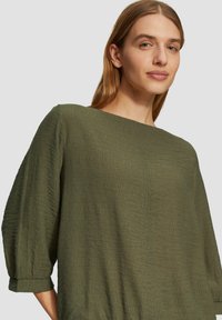 Blouse vert olive, texturée, à coupe ample, avec un décolleté bateau et des manches trois-quarts, présentant des détails de couture subtils.