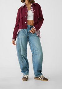 Bordeaux jakke med en stribet krave, parret med løse blå jeans med sømdetaljer. Snørebåndsbrune sneakers fuldender outfittet.