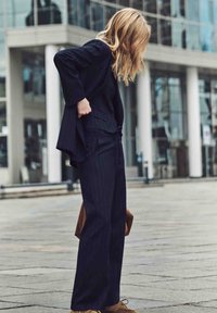 Costume sur mesure bleu marine à fines rayures avec un pantalon à jambes larges, tissu texturé et blazer ajusté, associé à des chaussures marron et un sac beige.