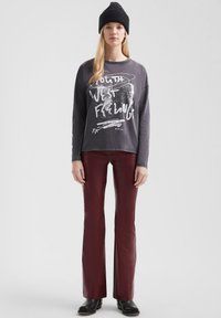 Grijze langemouwen shirt met witte grafische print, gecombineerd met bordeauxrode flared broek en zwarte enkellaarsjes. Donkere gebreide beanie op het hoofd.