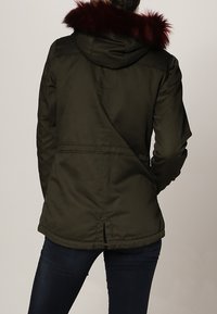 Osoba w ciemnozielonym parku z kapturem, wykończonym burgundowym futrem, w połączeniu z ciemnoniebieskimi dżinsami, stoi tyłem do widza.