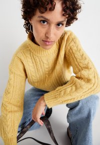 Pulover galben din tricot cu guler înalt și mâneci lungi, asortat cu blugi de culoare albastră deschisă. Puloverul are modele texturate.