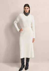Long ribbed knit dress in off-white, featuring a V-neck and long sleeves. Paired with a black geometric patterned scarf and black ankle boots.