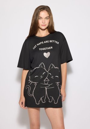 Giovane donna che indossa una camicia nera oversize con due gatti dei cartoni animati che si abbracciano e il testo "Cat naps are better together" con un cuore sopra.