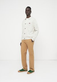 Ανοιχτό γκρι κουμπωτή ζακέτα με υφή. Καφέ κουμπιά και μικρό λογότυπο στο στήθος. Συνδυασμένο με μπεζ παντελόνι και πράσινα αθλητικά παπούτσια.