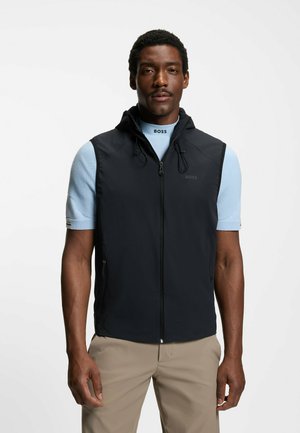 Mann trägt schwarze, zip-up Kapuzenweste über hellblauem Kurzarmshirt mit "BOSS"-Schriftzug und beiger Hose, steht vor weißem Hintergrund.