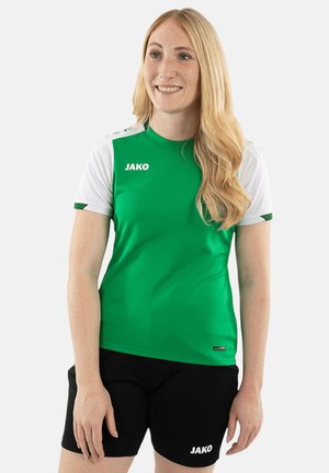 Donna con capelli biondi lunghi che indossa una maglietta sportiva JAKO verde e bianca e pantaloncini neri, sorridente e in piedi davanti a uno sfondo semplice.