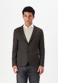 Donkergrijze blazer van textuurstof, met een enkelvoudige sluiting, twee voorzakken en twee knopen. Gedeeltelijk gedragen over een lichte blouse.