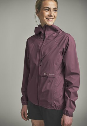 Woman smiling and wearing a purple hooded outdoor jacket with a front zipper and Ortovox logo, paired with black shorts.