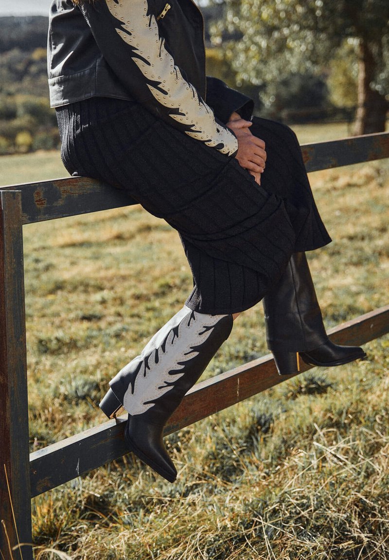 Knielange Stiefel mit schwarzem Lederdesign und weißem Schaft, verziert mit Blattmustern und Perforationen. Sitzt auf einem Zaun.