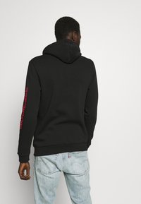 Sudadera negra con capucha y puños de canalé, que presenta texto rojo en la manga derecha. Combinada con jeans azul claro. Textura de tela suave.