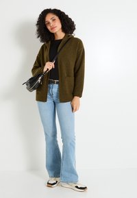 Cardigan texturé vert olive à manches longues, devant ouvert et deux poches, associé à un jean évasé bleu clair et des baskets multicolores.