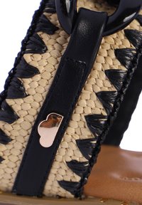 Sandalias trenzadas con un diseño en beige y negro, que cuentan con una parte superior trenzada, una correa de cuero y un acento metálico. Suela plana.
