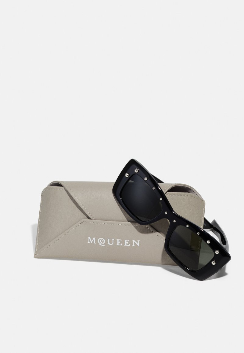 Occhiali da sole neri oversize con accenti di borchie argentate, poggiati su un elegante astuccio protettivo beige chiaro impresso con la scritta "MQUEEN."