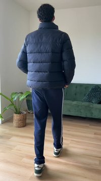 Fekete puffermellény vízszintes steppelt mintával, kék sportnadrággal, fehér csíkkal, és zöld árnyalatú sportcipővel párosítva.