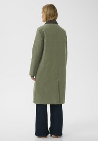 Olijfgroen lange jas met een donkere kraag, met een rechte snit en zijsplitten; gedragen over donkere wijde jeans en tan schoenen.