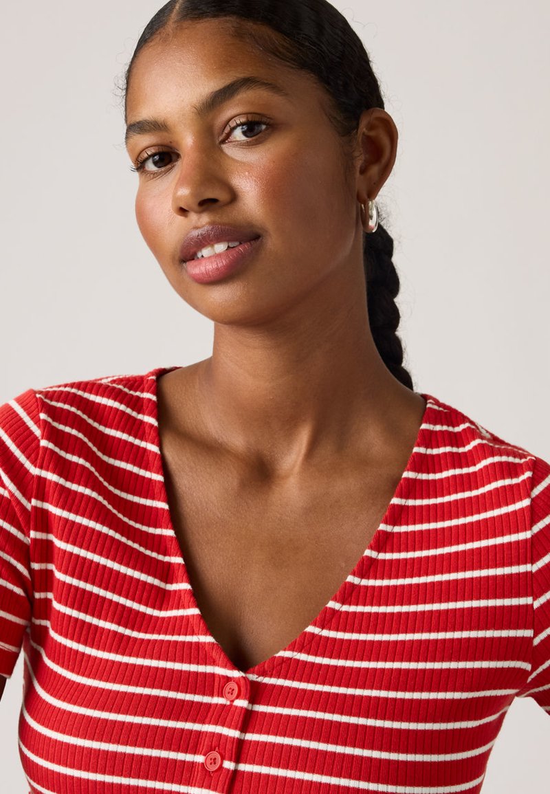 Jeune femme aux cheveux tressés portant des boucles d'oreilles argentées en forme de créoles et un t-shirt à col en V côtelé rouge et blanc à rayures avec des boutons.