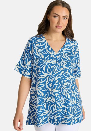 Vrouw draagt een blauw kortemouwenblouse met een wit abstract bladerenpatroon, V-hals en losse pasvorm, gecombineerd met een witte broek.