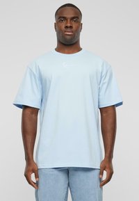 Light blue cotton T-shirt with short sleeves, a crew neckline, and a small white logo on the left chest, worn with light denim jeans.