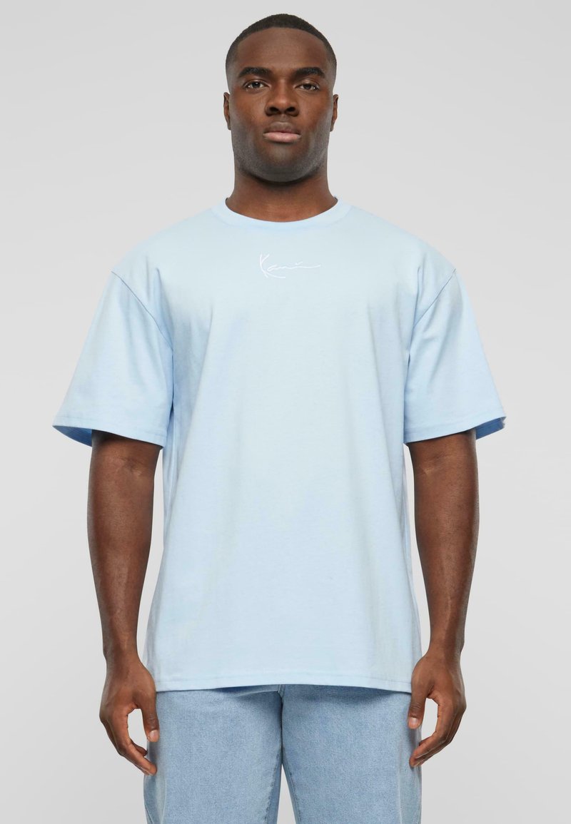 Light blue cotton T-shirt with short sleeves, a crew neckline, and a small white logo on the left chest, worn with light denim jeans.