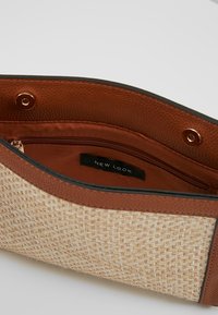 Woven tote bag featuring a beige body with gold threads, brown leather accents, and an interior zip pocket with a black label.