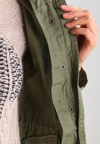 Olivgrüne Jacke mit Metall-Druckknöpfen und strukturiertem Futter, geschichtet über einem leichten gestrickten Pullover mit schwarz-weißen Mustern.