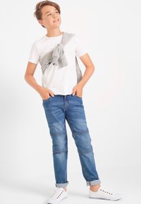 Maglietta bianca, maglione grigio chiaro annodato sulle spalle, jeans in denim blu con un lavaggio chiaro, orli arrotolati e sneakers bianche.