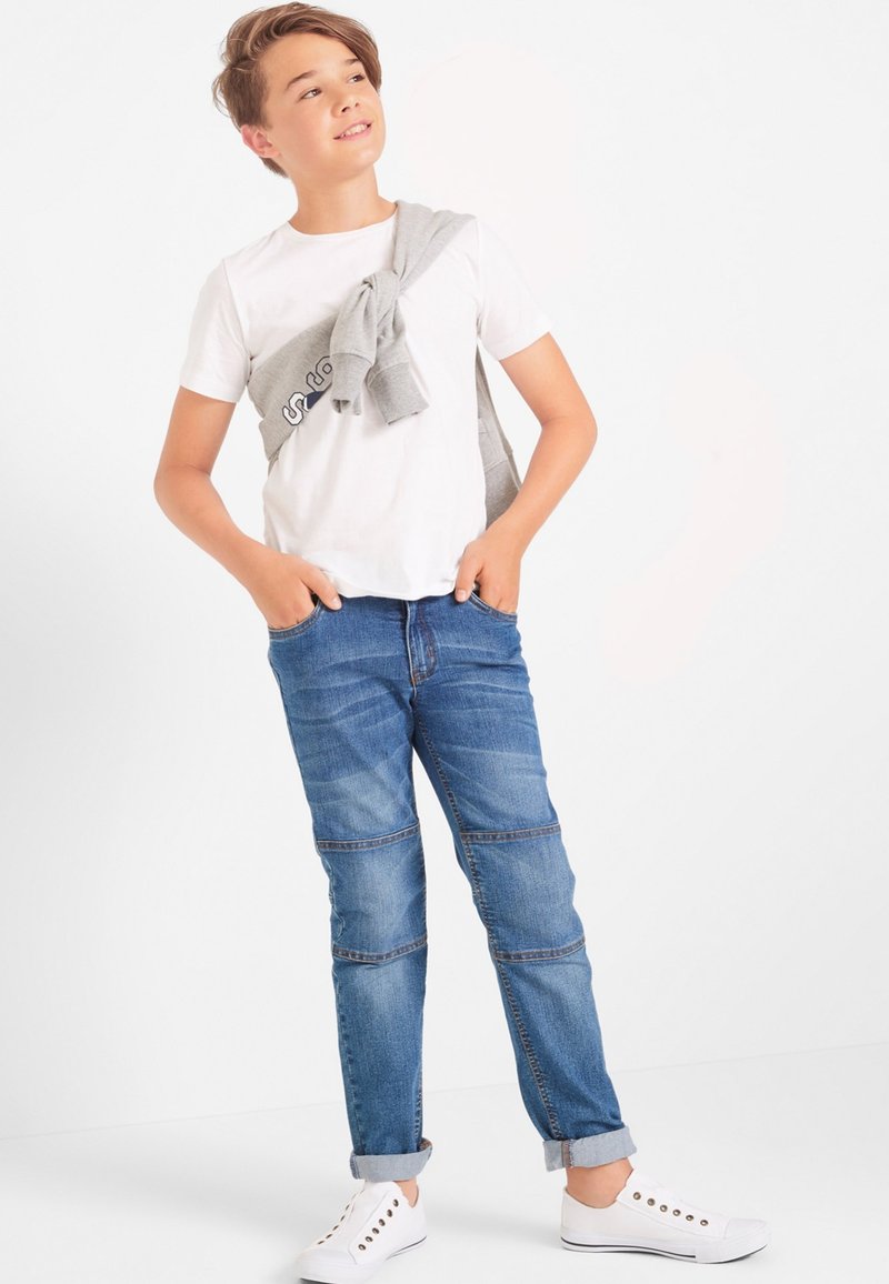 Maglietta bianca, maglione grigio chiaro annodato sulle spalle, jeans in denim blu con un lavaggio chiaro, orli arrotolati e sneakers bianche.