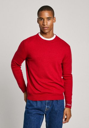 Suéter rojo de cuello redondo hecho de un material de punto suave. Presenta puños acanalados, un cuello blanco en contraste y un logo sutil en la manga.