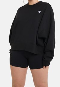 Černá oversize mikina se žebrovaným lemem, která má malé bílé logo na levé části hrudi, doplněná černými žebrovanými šortkami.
