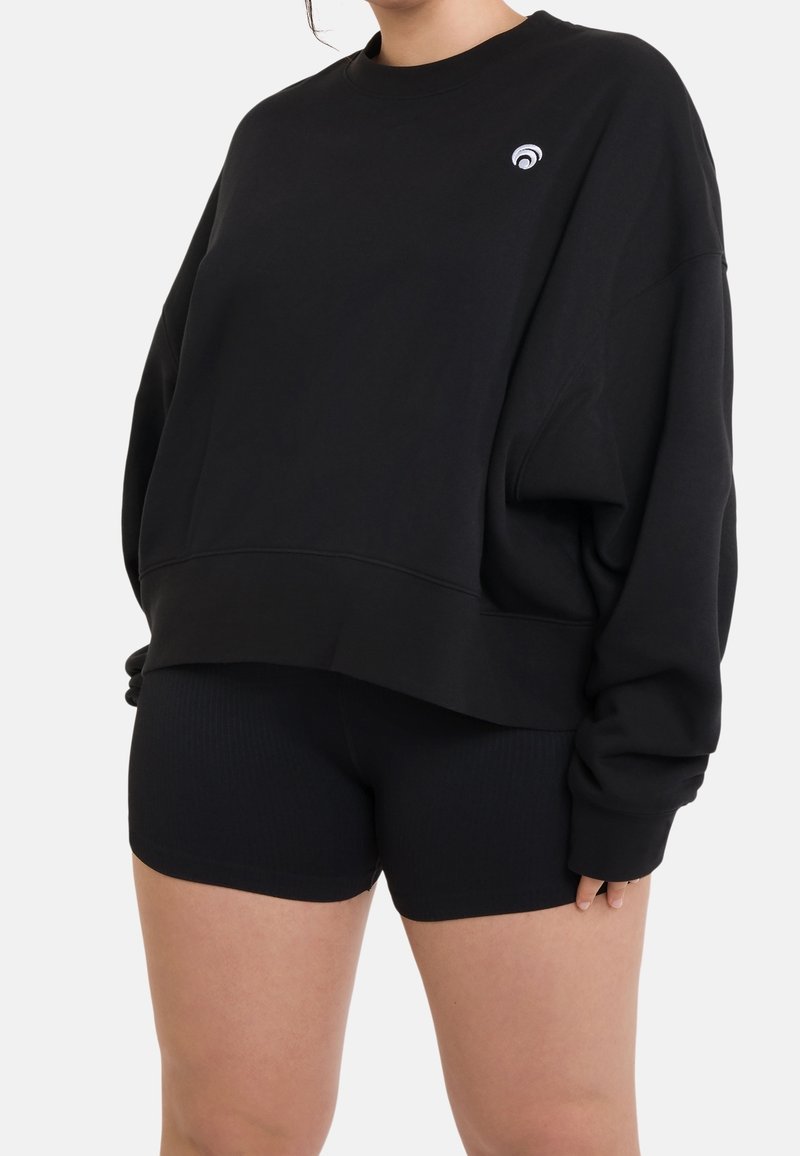 Černá oversize mikina se žebrovaným lemem, která má malé bílé logo na levé části hrudi, doplněná černými žebrovanými šortkami.