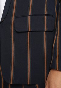 Granatowa marynarka w pionowe paski w kolorze rdzy, z wyraźnym detalem tekstury i kieszenią na piersi, świadcząca o dopasowanym kroju.