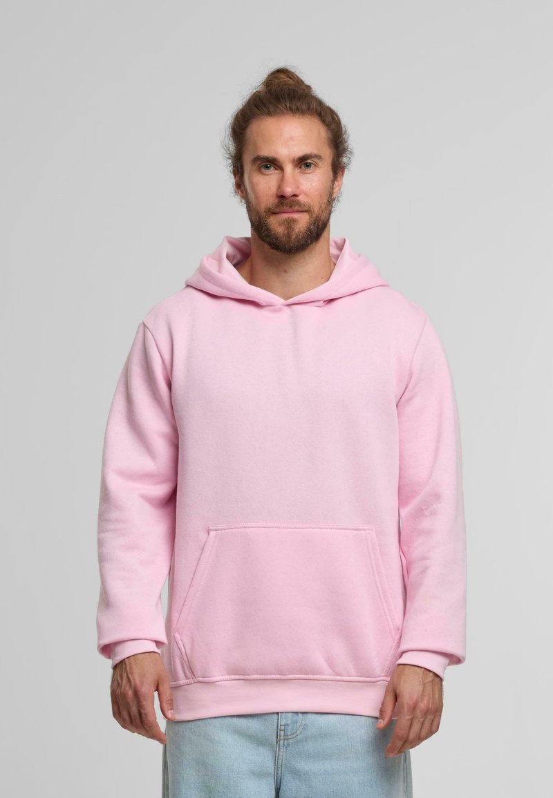 Sudadera rosa hecha de un tejido suave, presenta un bolsillo frontal, una gran capucha y puños acanalados. Llevada sobre unos jeans de color azul claro.