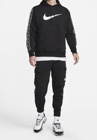 Mann trägt schwarzen Nike-Hoodie und schwarze Cargo-Jogginghosen mit weißen Socken und schwarz-weißen Nike-Sneakern, steht vor weißem Hintergrund.