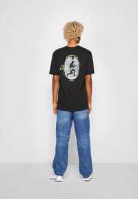 Svart T-skjorte med grafisk design som viser en figur og tekst på baksiden; kombinert med blå denim vide bukser og hvite sneakers.
