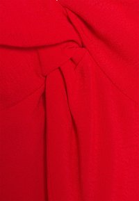 Červená pletená textilie s nabíranými sekcemi a detailním provázáním na rameni, což vytváří strukturu a vizuální zajímavost.